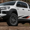 White Toyota Hilux GR Sport fitted with premium multi-spoke aftermarket 4x4 alloy wheels, parked on a rugged dirt road with rocky mountain terrain in the background.
